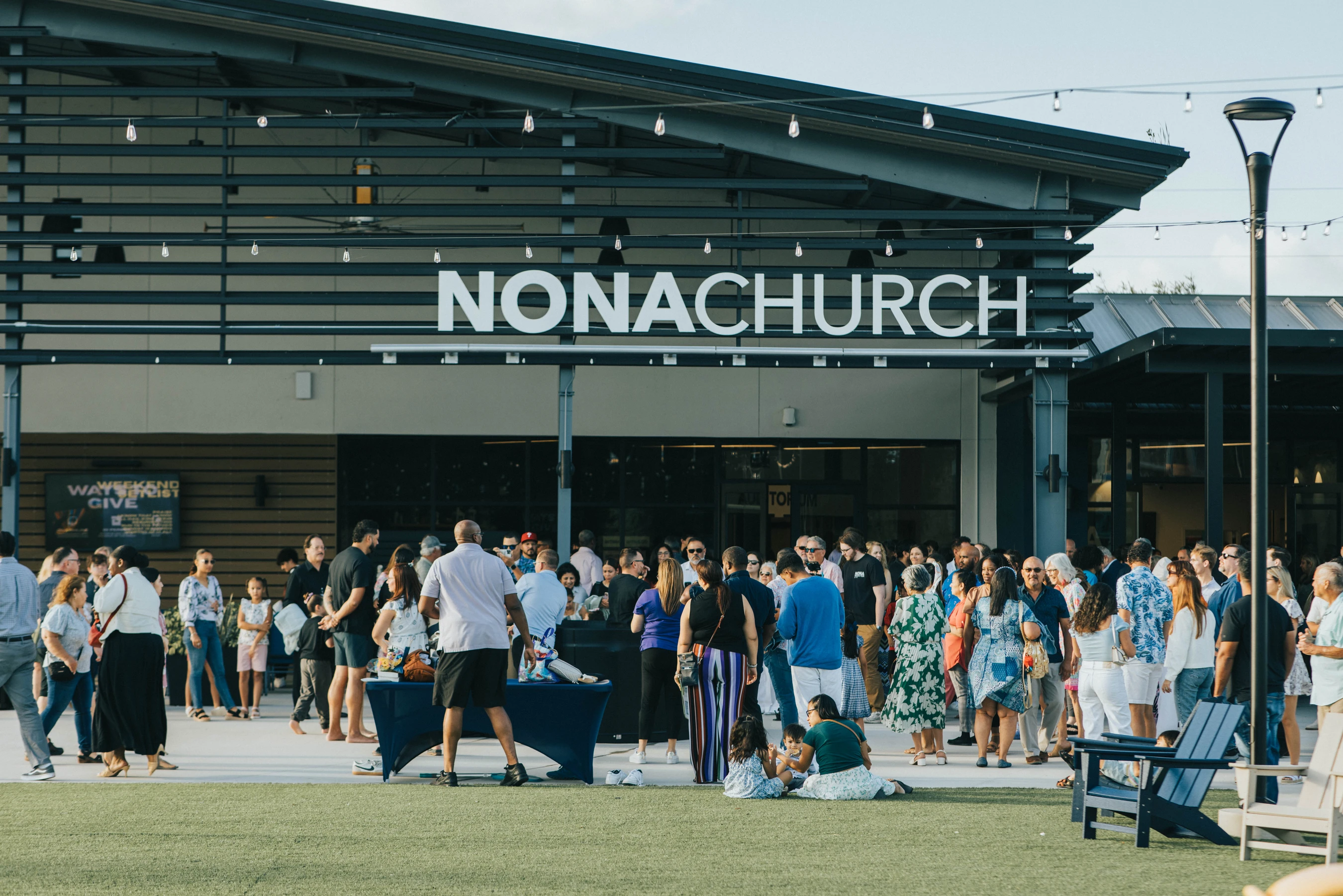 A diverse crowd of people socializes on a patio and lawn outside a modern building with a large white 'NONACHURCH' sign under hanging string lights.