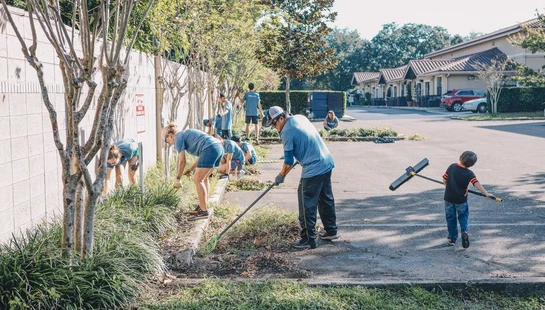 A group of adult volunteers and a young boy clean up a landscaped area along a wall and parking lot, weeding, raking, and sweeping on a sunny day.