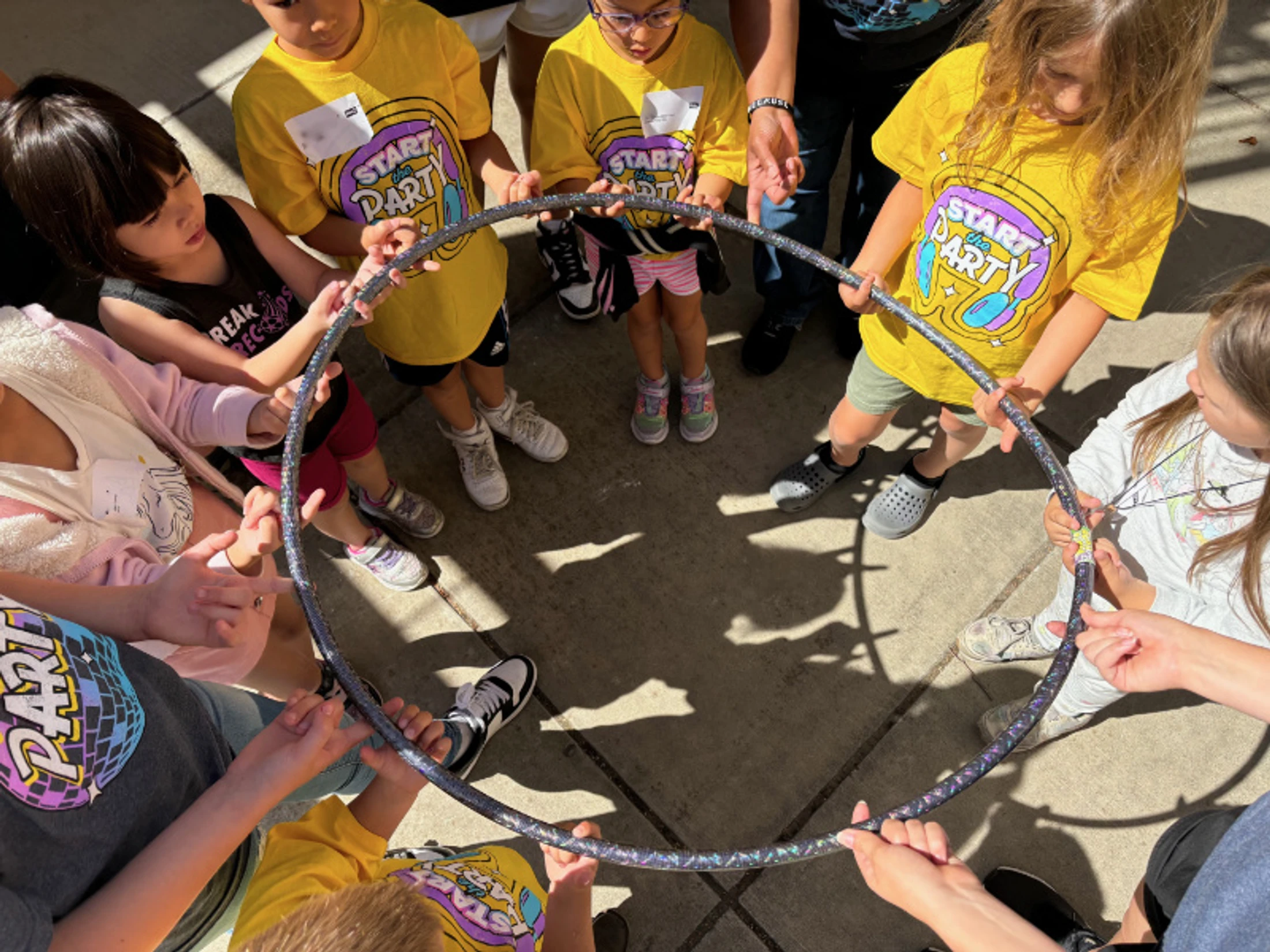 An overhead view of a group of children standing in a circle, all holding onto a hula hoop together as part of a team-building activity.