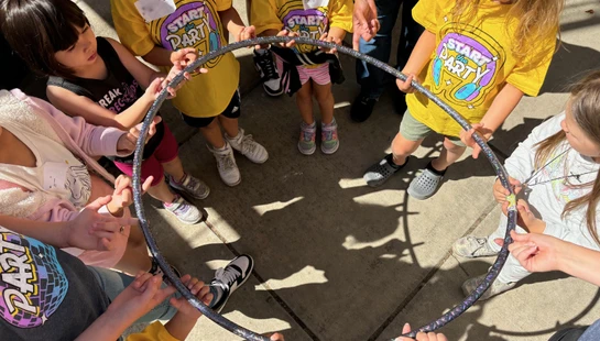 An overhead view of a group of children standing in a circle, all holding onto a hula hoop together as part of a team-building activity.