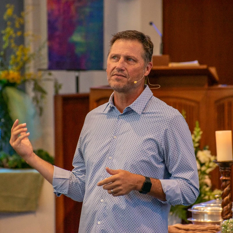 A middle-aged man in a blue patterned shirt with a microphone on his face speaks and gestures while standing in a church with a pulpit in the background.