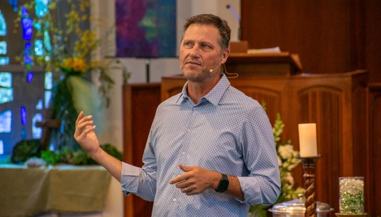 A middle-aged man in a blue patterned shirt with a microphone on his face speaks and gestures while standing in a church with a pulpit in the background.
