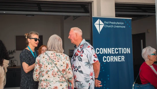 A group of people chat together outdoors in front of a blue banner for the First Presbyterian Church Livermore that says 'Connection Corner'.