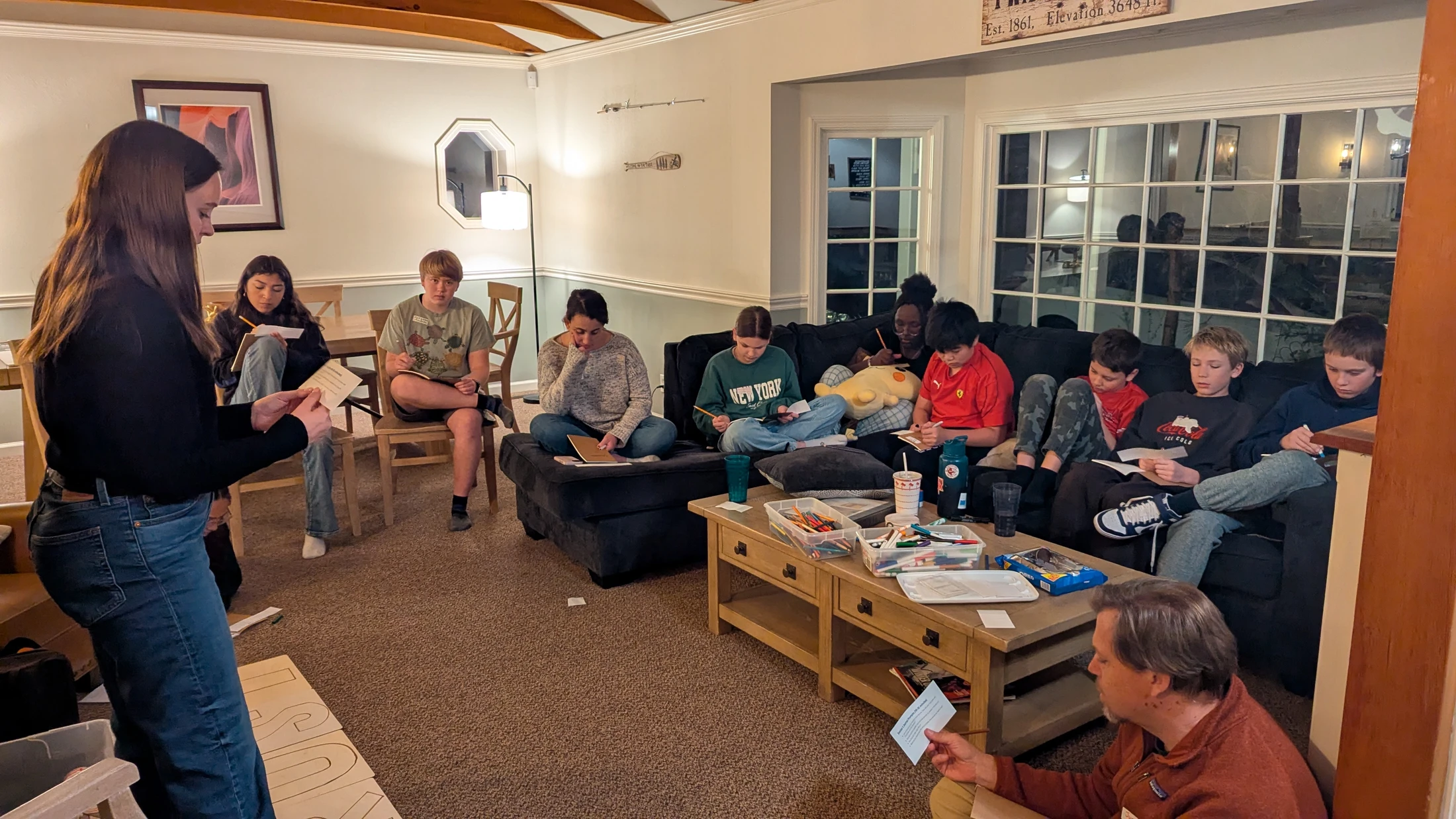 A group of teenagers and adults sit in a living room, focused on reading and writing on small cards during a quiet, organized activity.