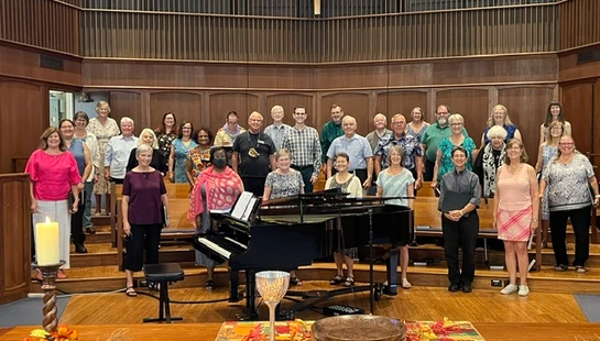 A large, diverse choir of adults stands on risers in a church sanctuary, smiling for a group photo in front of a grand piano and a large pipe organ.
