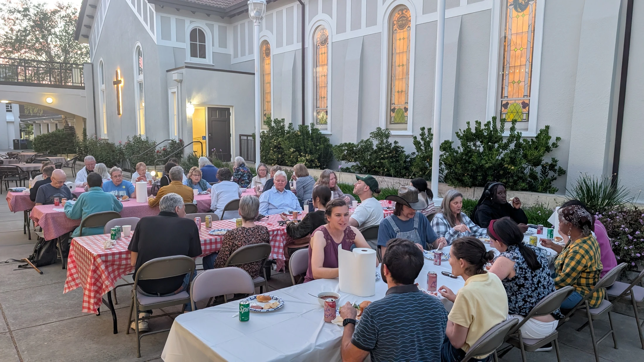 A diverse group of people sit at long tables covered in tablecloths, eating and socializing in the courtyard of a church building at dusk.
