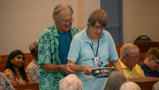 An older man in a green patterned shirt assists another man holding a metal collection plate as they walk through pews in a church or meeting hall.