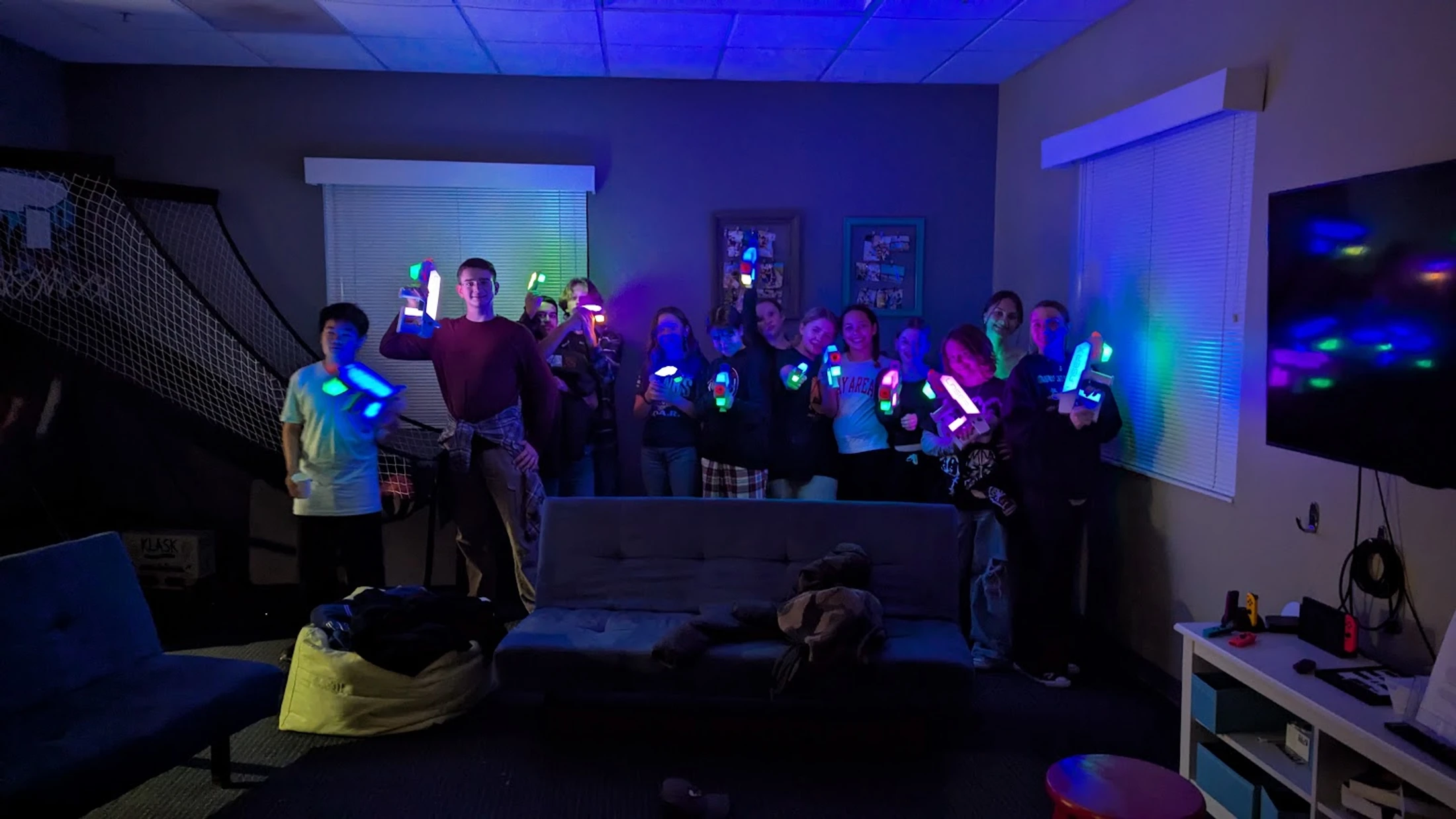A group of young people posing in a dark room illuminated by blue light, holding colorful glowing laser tag guns and smiling for the camera.