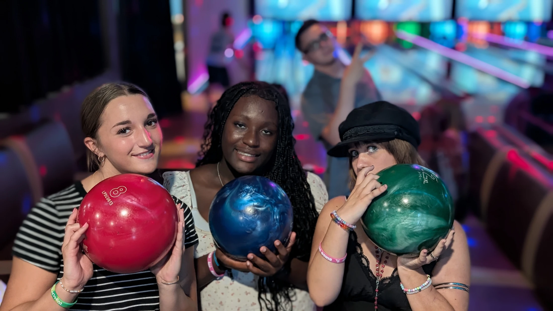 Three young women smile and pose for the camera, each holding a colorful bowling ball in a neon-lit bowling alley. A man is visible in the background.