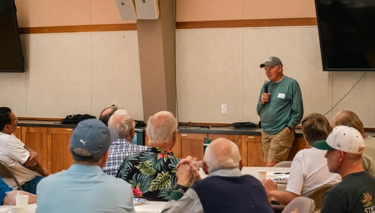 A man in a green shirt and baseball cap speaks into a microphone, addressing a seated audience of mostly older men in a meeting room.