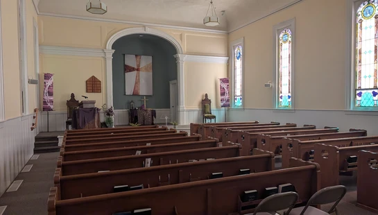The empty interior of a sunlit church sanctuary, showing rows of wooden pews facing an altar with a pulpit and a cross tapestry under an archway.
