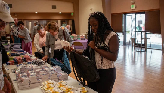A smiling young woman with long braids packs a backpack with free school supplies from a table, while other community members select items at an indoor event.