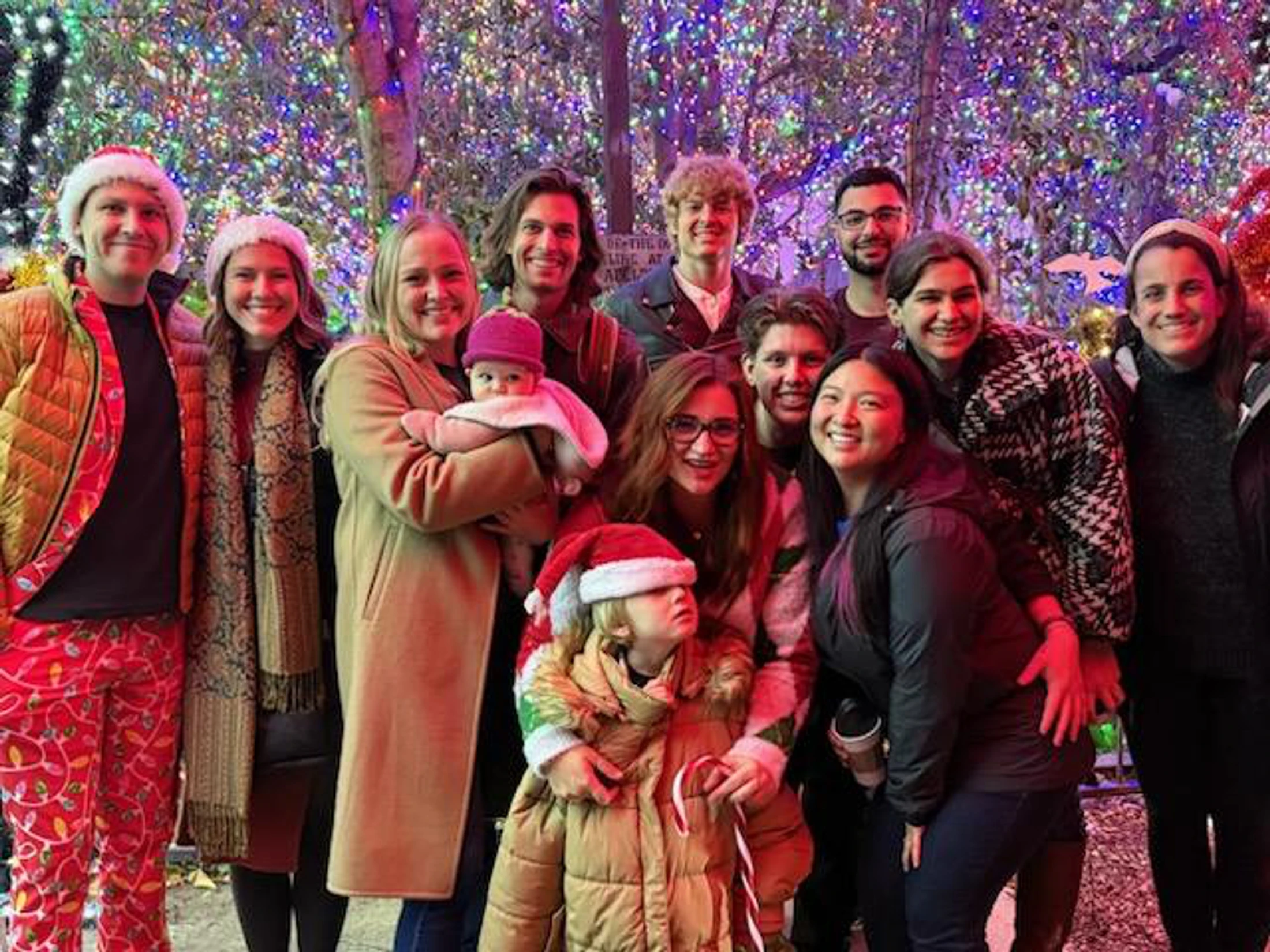 A large group of smiling friends and family, including a child and a baby, pose for a photo at night in front of trees covered in colorful Christmas lights.