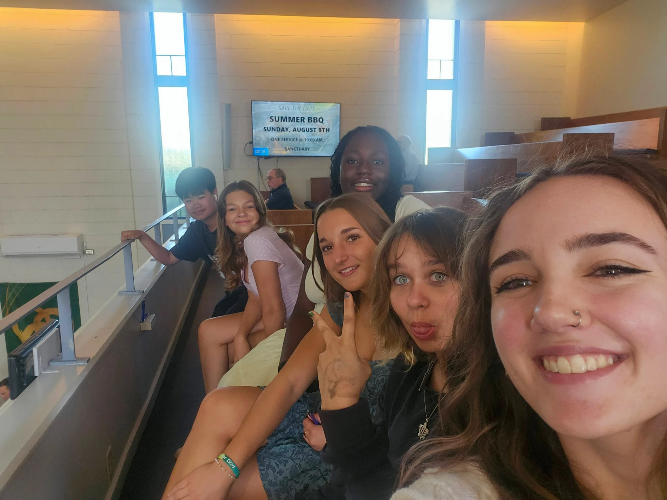 A smiling young woman takes a selfie with a diverse group of five friends sitting behind her in the tiered seating of a church or auditorium.
