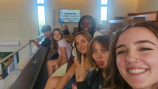A smiling young woman takes a selfie with a diverse group of five friends sitting behind her in the tiered seating of a church or auditorium.