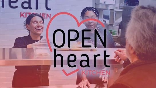 Two smiling women volunteers serve food on a tray to a man at a soup kitchen counter, with an 'Open Heart Kitchen' logo in the foreground.