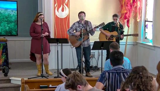 A band of three musicians, a woman singing and two men playing acoustic guitars, performs on stage for an audience seated in pews in a sunlit church.
