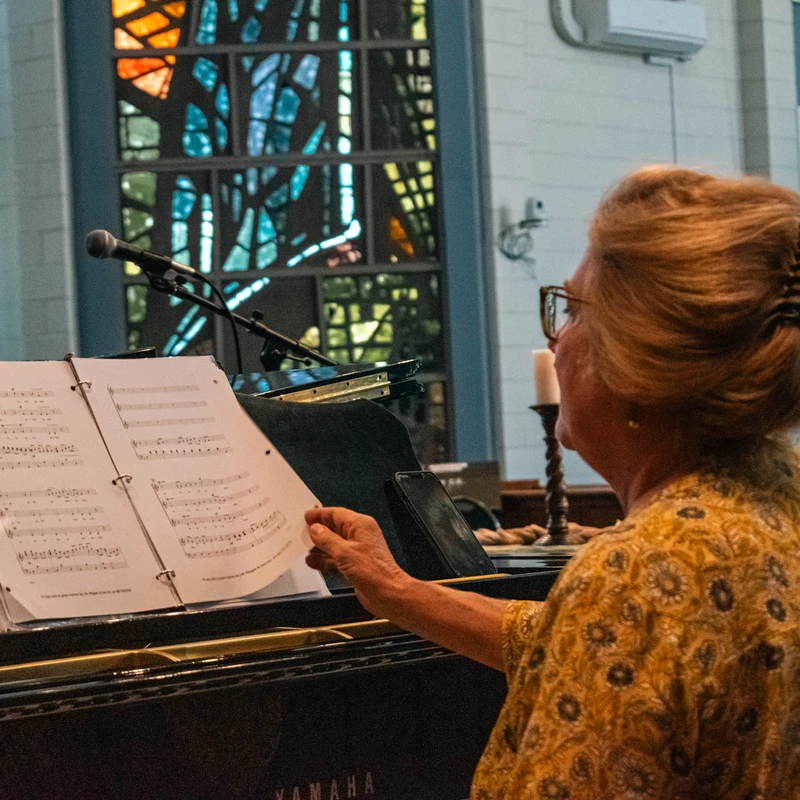 A woman seen from behind sits at a piano in a church, turning the page of sheet music. A colorful stained-glass window is visible in the background.