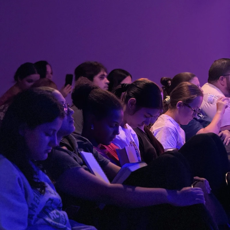 A diverse group of young people sit in a dark room, illuminated by purple light. Many are looking down, focused on notebooks they hold in their laps.