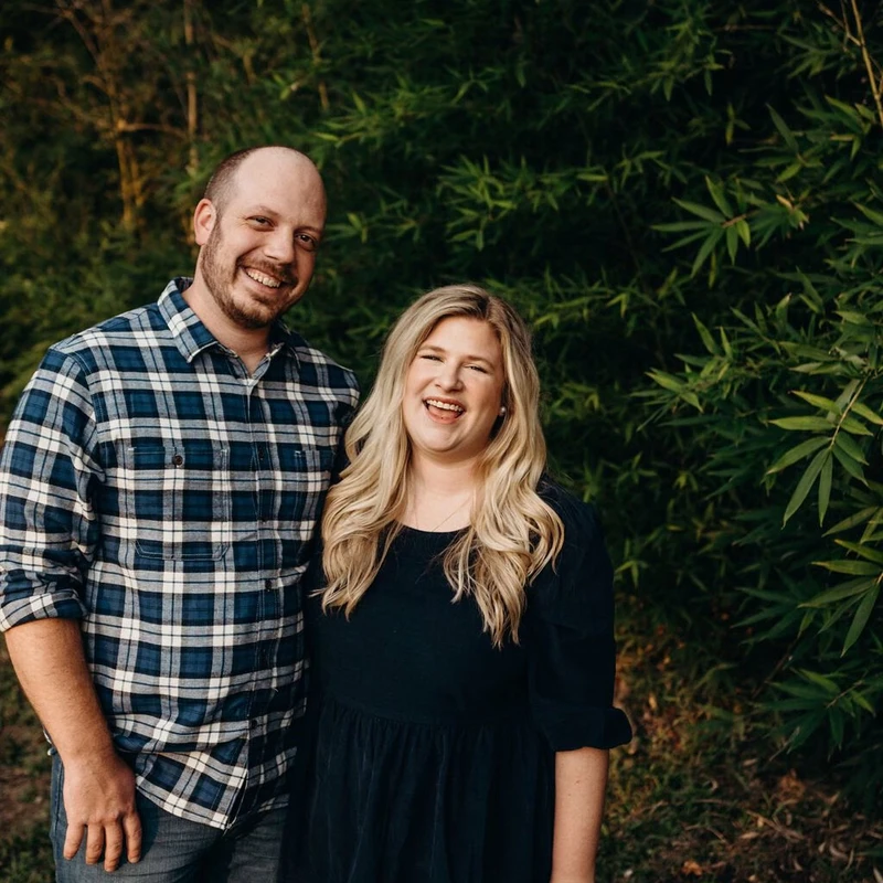 A smiling man in a plaid shirt and a laughing woman in a dark dress stand together outside in front of a wall of green bamboo leaves.