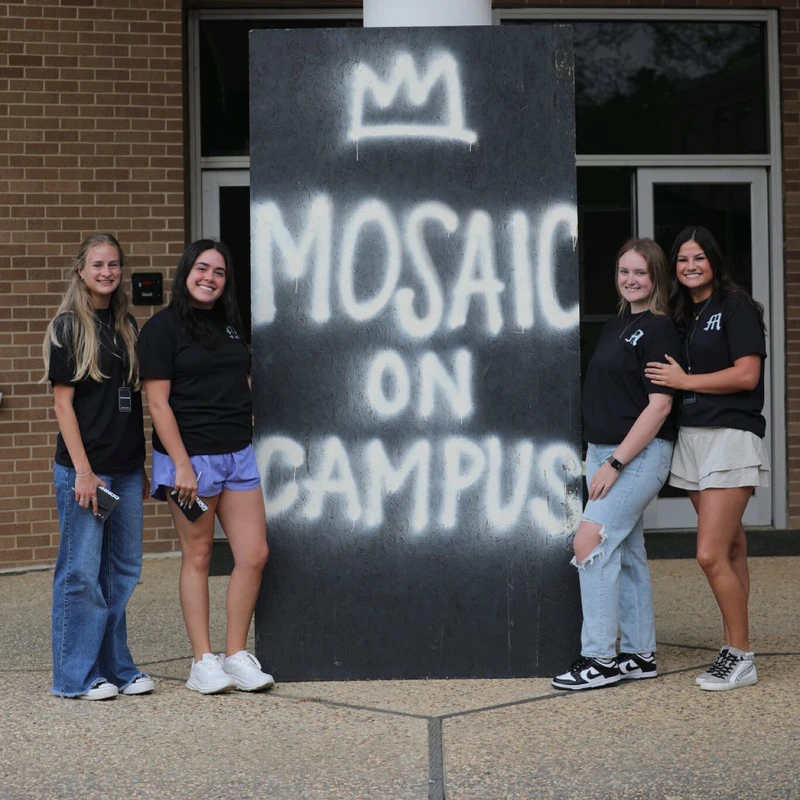 Four young women smile for a photo, posing on either side of a large black sign with 'MOSAIC ON CAMPUS' spray-painted in white.