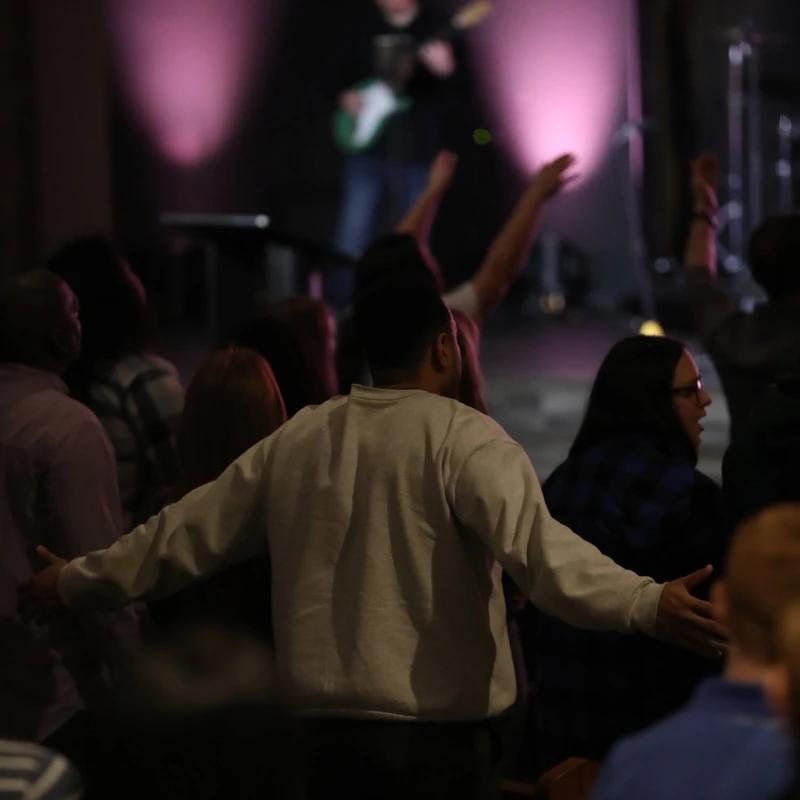 A man in a grey sweatshirt stands with arms outstretched in a crowd of people worshiping. A musician is on the dimly lit stage in the background.