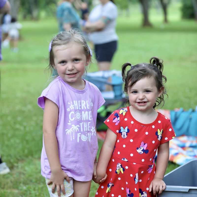 Two young girls stand close together in a grassy park, smiling at the camera. One wears a purple shirt, and the other wears a red Minnie Mouse dress.