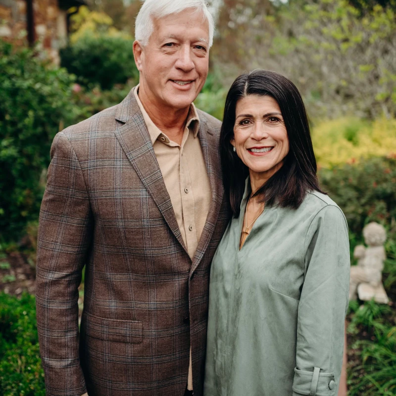 A man with white hair in a plaid blazer and a woman with dark hair in a green dress stand close together and smile at the camera in a lush garden.