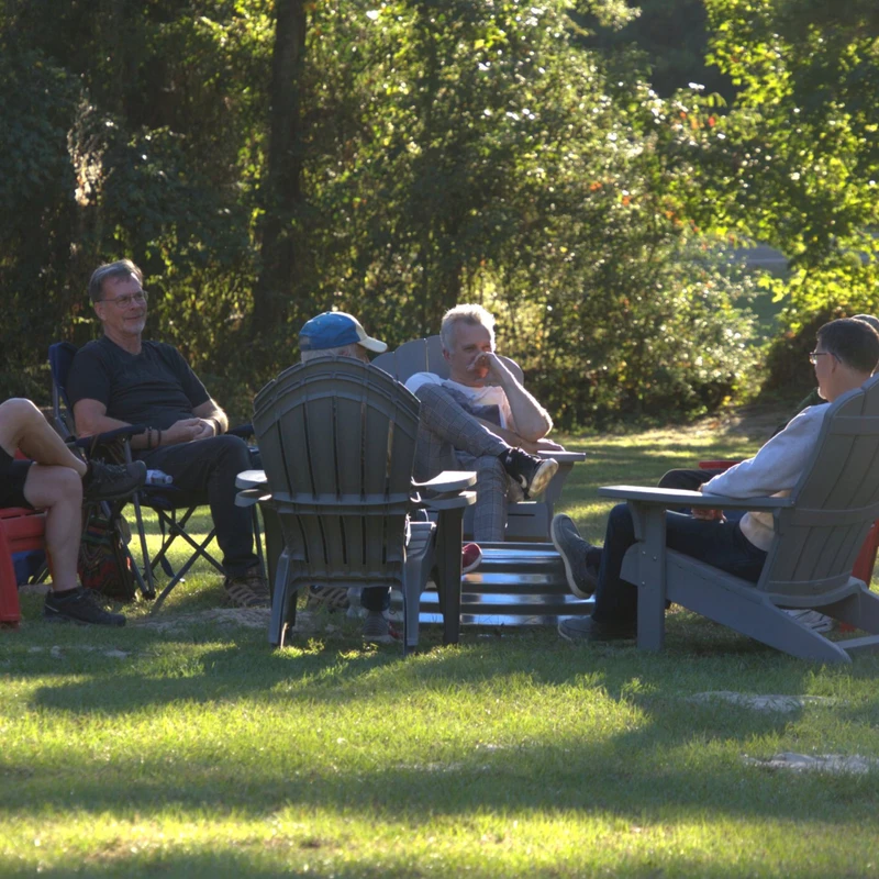 A group of six men sit in a circle of lawn chairs on a sunny, grassy area, engaged in conversation with trees in the background.