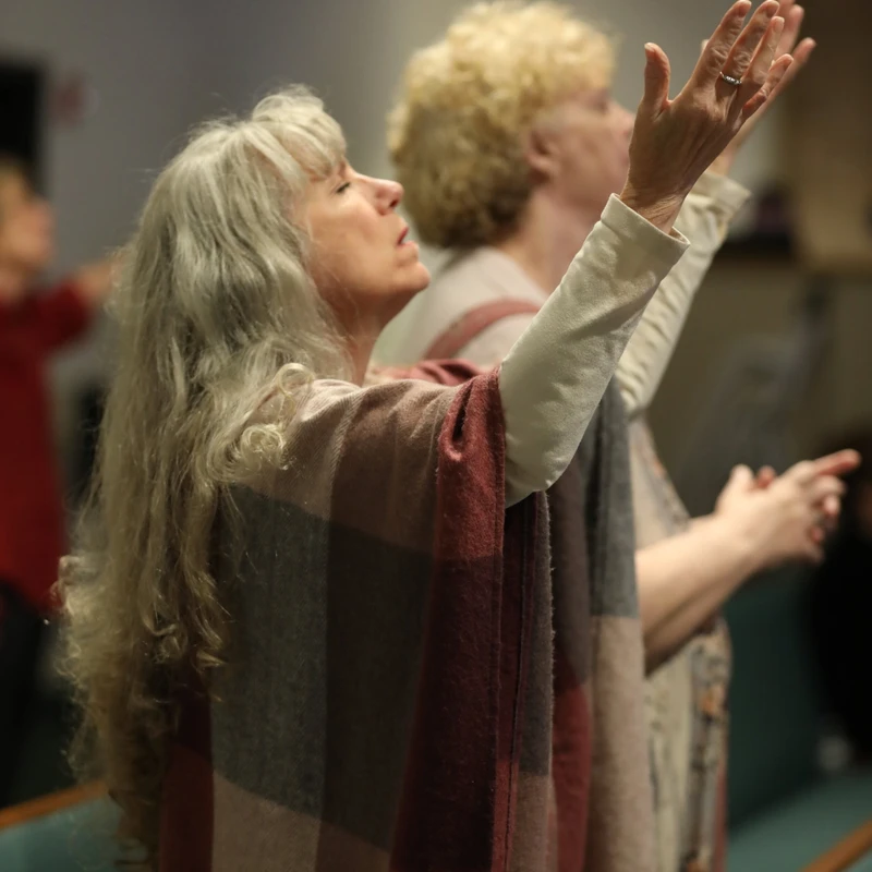 A woman with long grey hair wearing a plaid shawl lifts her hands in worship during a church service, with her eyes closed and head tilted back.