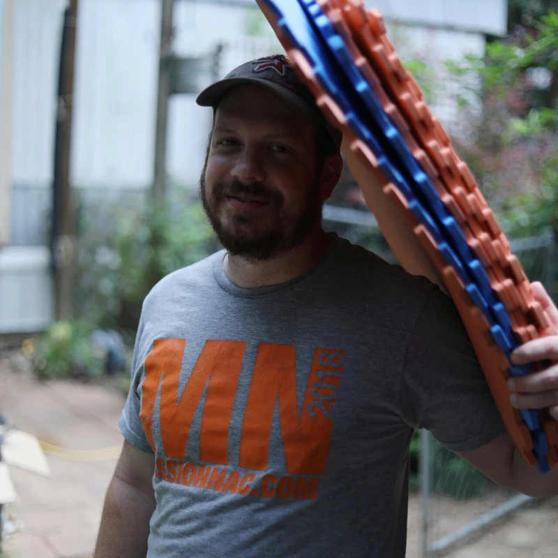 A smiling man with a beard and a baseball cap carries a stack of orange and blue interlocking foam floor mats over his shoulder in a backyard.