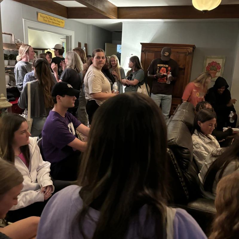 A large group of young adults are gathered in a crowded living room with wood beams, talking and socializing. Some stand while others sit on a couch.