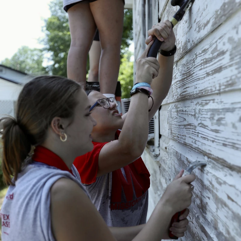 A group of young volunteers uses scrapers and hammers to remove old, flaking white paint from the wooden siding of a house on a sunny day.