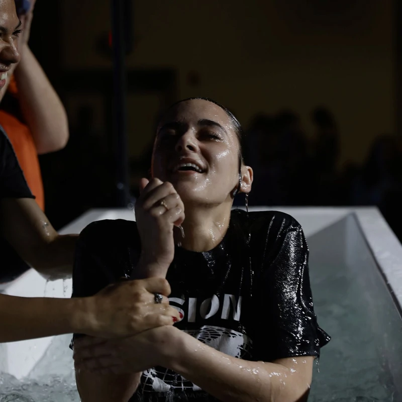 A young woman with wet hair and a black t-shirt emerges from a baptismal pool with her eyes closed and a peaceful smile, being assisted by another person.