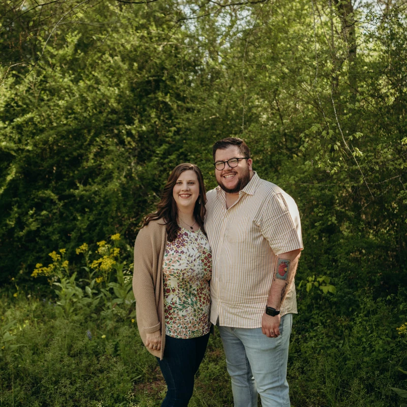 A smiling man and woman stand close together for a portrait in a grassy area, with a dense green forest and wildflowers in the background.