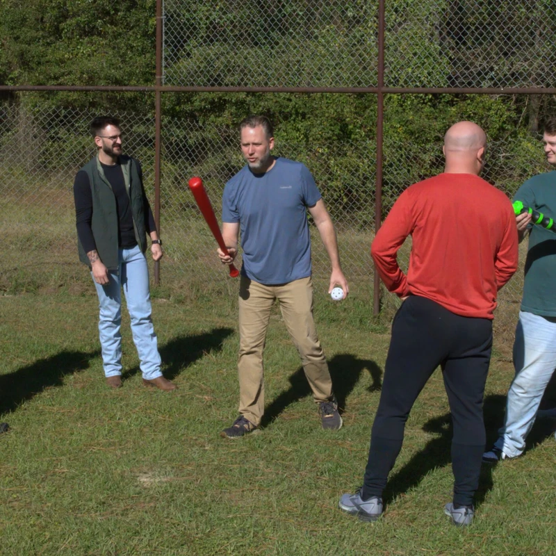 A group of eight men in casual clothes stand in a grassy field with a chain-link fence behind them, some holding wiffle ball bats, preparing to play a game.