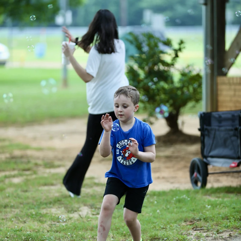 A young boy in a blue shirt walks on grass, looking intently at soap bubbles floating around him. A woman is blurred in the background.
