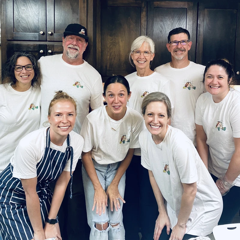 A group of eight smiling adults pose for a photo in a kitchen with dark wood cabinets. They are wearing matching white t-shirts with a small cartoon logo.