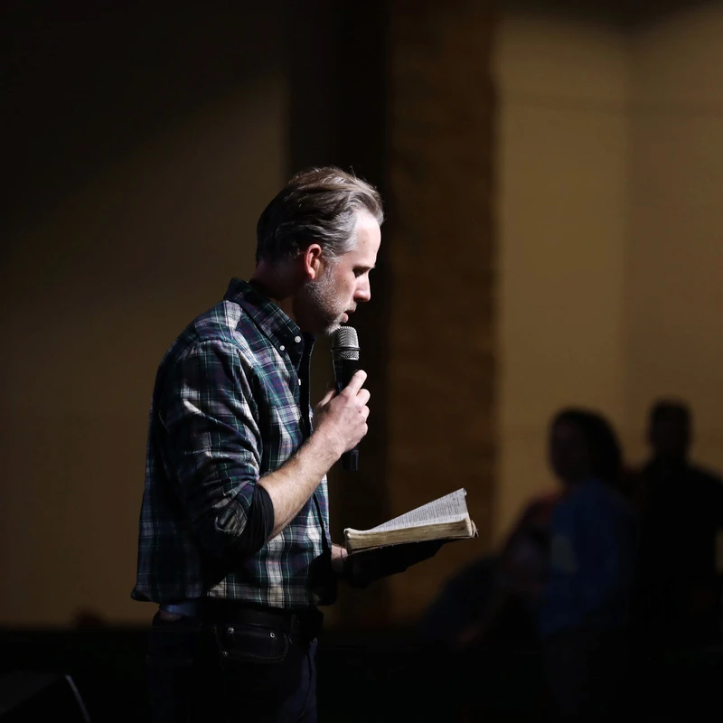 A man in a plaid shirt stands in a spotlight on a dark stage, reading from a book into a microphone. A blurry audience is in the background.