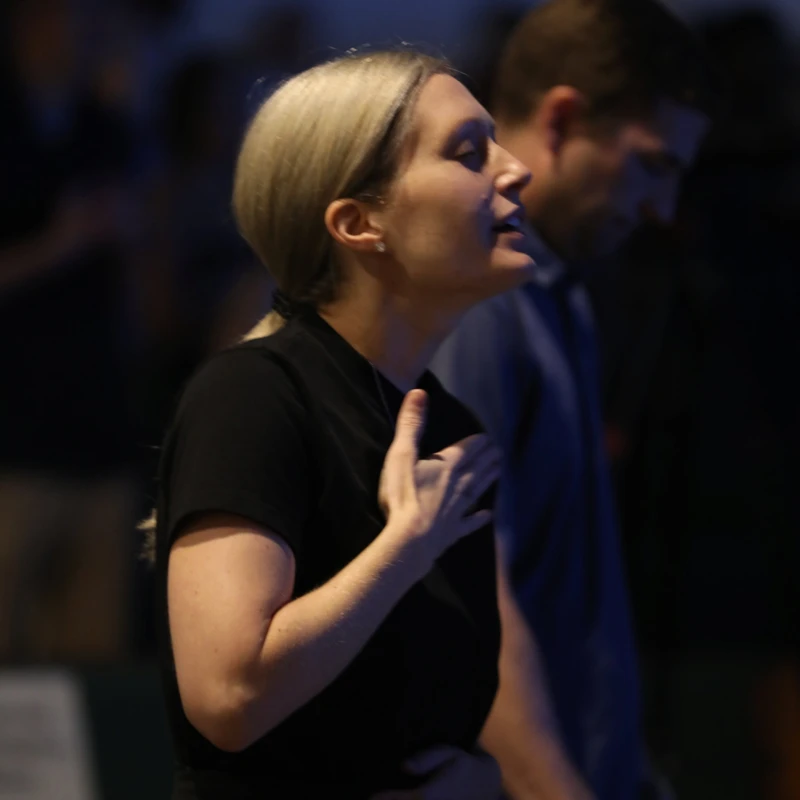 A blonde woman in a black shirt stands with her eyes closed and a hand over her heart, appearing to be in a moment of deep worship or prayer in a dim setting.
