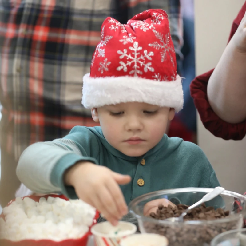 A young boy wearing a red Santa hat with snowflakes on it reaches for toppings at a holiday treat station with bowls of marshmallows and chocolate chips.