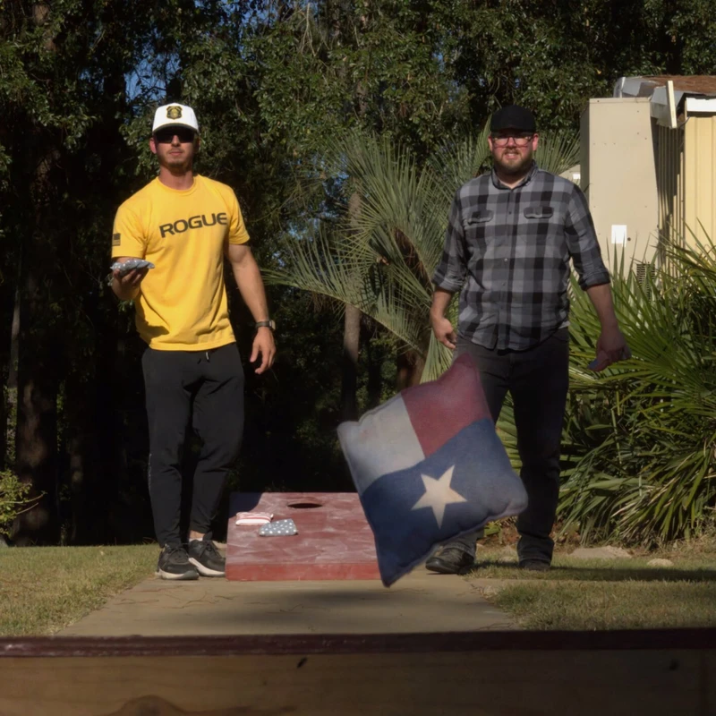 Two men play cornhole in a grassy yard. One man throws a beanbag with the Texas flag on it, captured in mid-air as it flies towards the board.