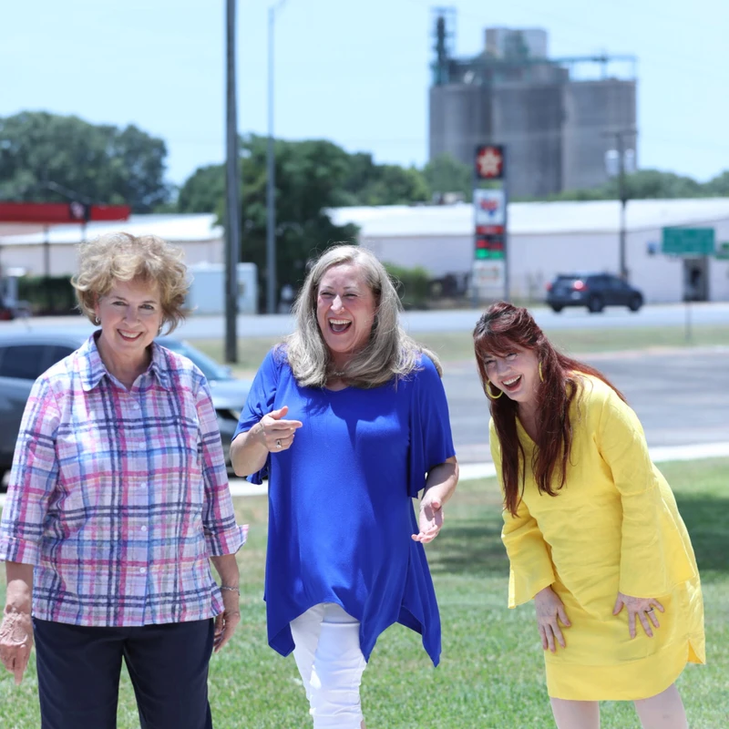 Three women stand on a sidewalk next to a lawn on a sunny day. Two are laughing heartily while the third smiles at the camera.
