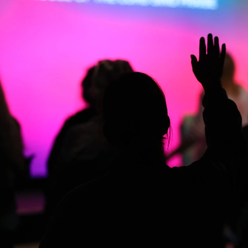 A silhouette of a person from behind with their right hand raised, against a bright pink and blue out-of-focus background screen at a church service.