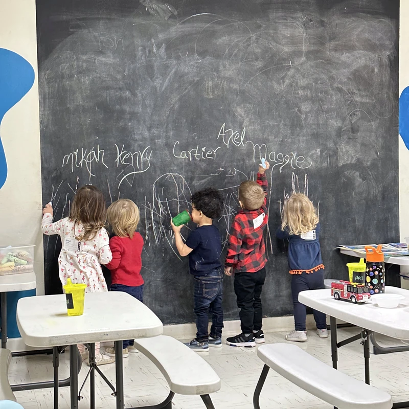 Five young children stand with their backs to the camera, drawing with chalk on a large blackboard that takes up most of the wall in a classroom setting.