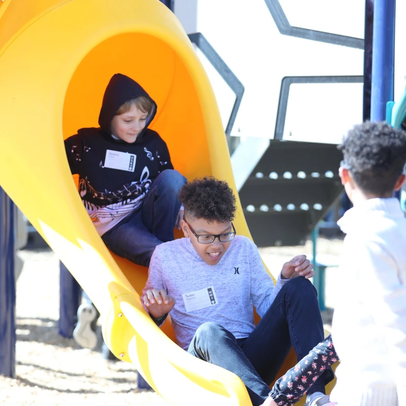 Two young boys slide down a bright yellow playground slide together on a sunny day, while other children play at the bottom.