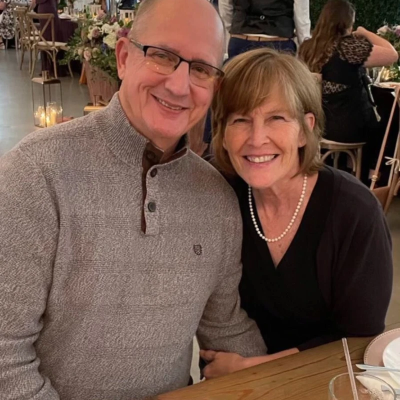 A man in a grey sweater and glasses and a woman in a black top with a pearl necklace smile for the camera while seated at a wooden table at a reception.