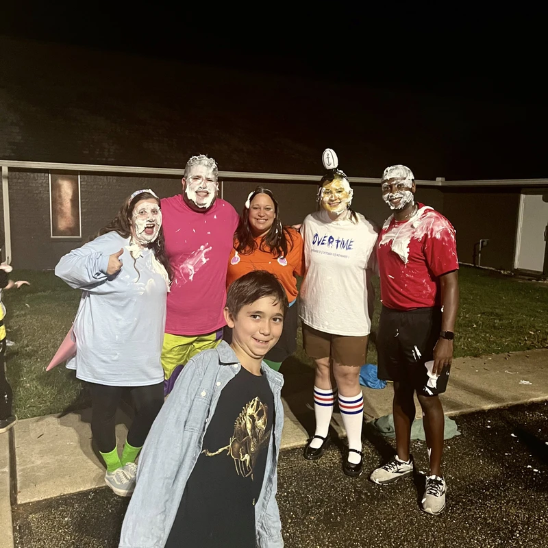 A young boy smiles in the foreground while five adults stand behind him, their faces and clothes covered in white cream after a fun game at night.