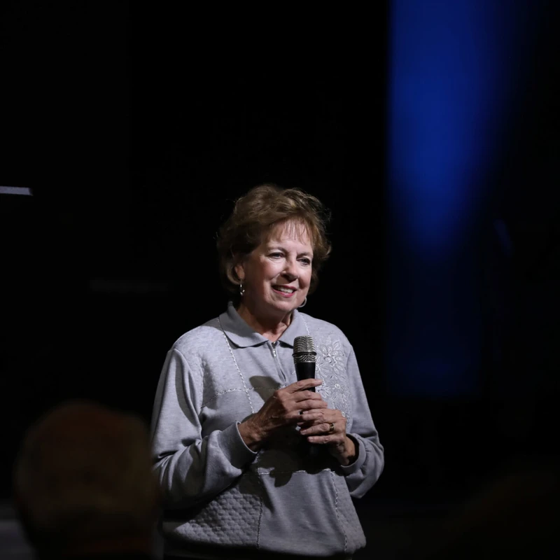 An older woman with short brown hair smiles while holding a microphone. She is on a dark stage, lit by a spotlight and a blue light in the background.