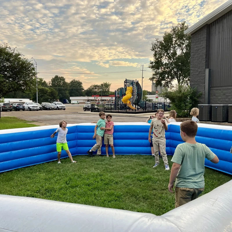 A group of young children plays inside a large, rectangular, blue and white inflatable arena set up on a grassy lawn under a cloudy evening sky.